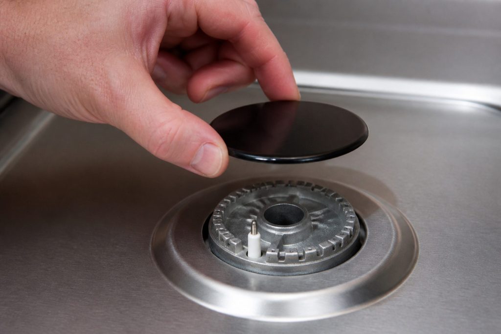 A hand lifting the enameled cap of a gas burner straight up off its head, an inch of air under it, the flame ports and the slim ignition electrode exposed in the well below.