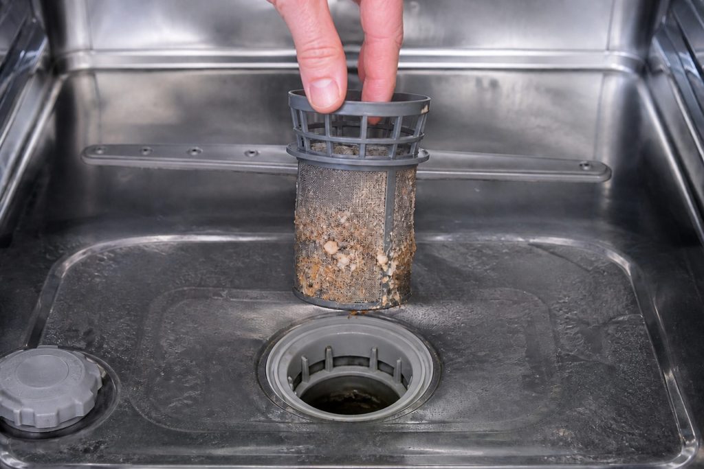 The cylindrical filter of a dishwasher twisted out and lifted straight up on two fingers, food debris matted around its mesh, the empty round sump opening below it in the tub floor.