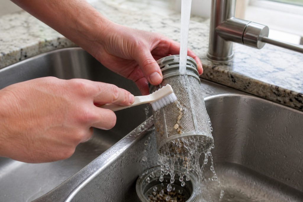 A dishwasher filter cylinder held under a running kitchen tap in one hand, water breaking through the mesh and food debris rinsing away down the sink, the other hand working a soft brush over it.