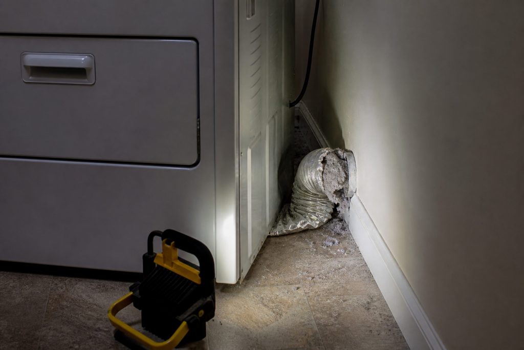 A dryer pulled two feet out from a laundry wall, seen from the side, the crushed foil duct behind it kinked flat against the baseboard and a grey wad of lint packed at the elbow where it enters the wall.