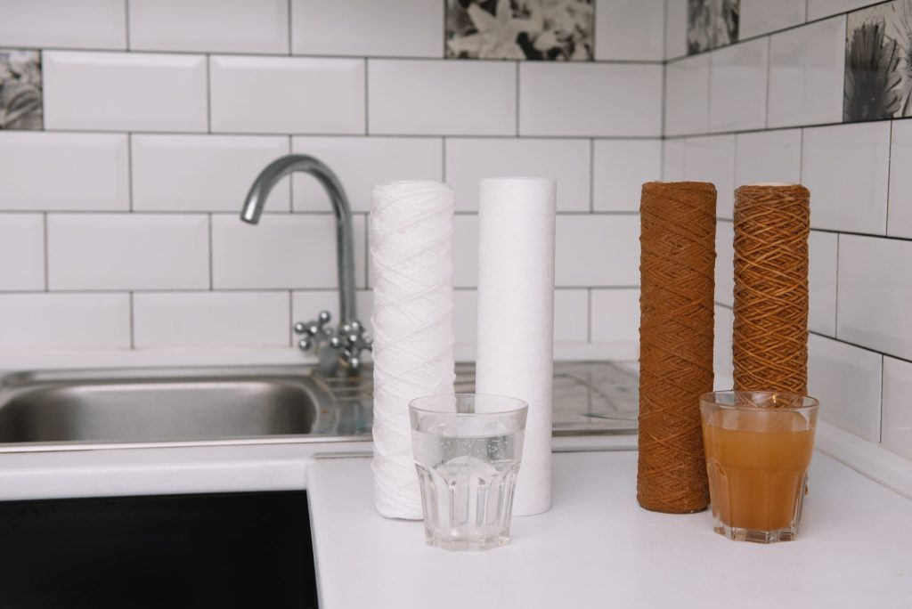 Two new white filter cartridges beside two brown exhausted ones on a kitchen counter, with a glass of clear water and a glass of dirty water