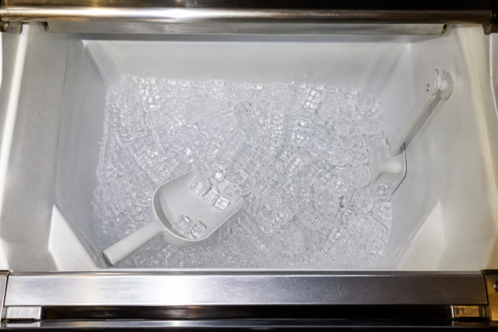 Looking straight down into the open storage bin of an undercounter ice machine from directly above, a shallow bed of clear cubes across the bottom, the plastic bin walls and the bin sensor arm at one side, the scoop half buried.