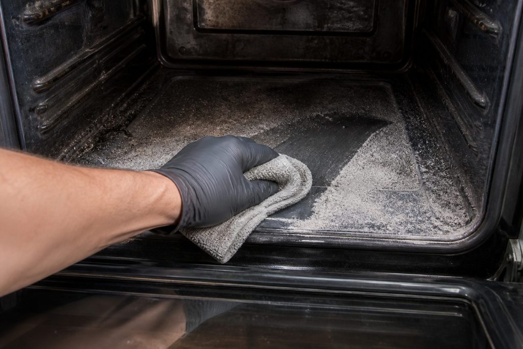 A gloved hand sweeping a damp cloth through white ash across the floor of an oven cavity after a high-heat cycle, the interior walls discolored and the racks pulled out, shot low from just inside the open door looking in.