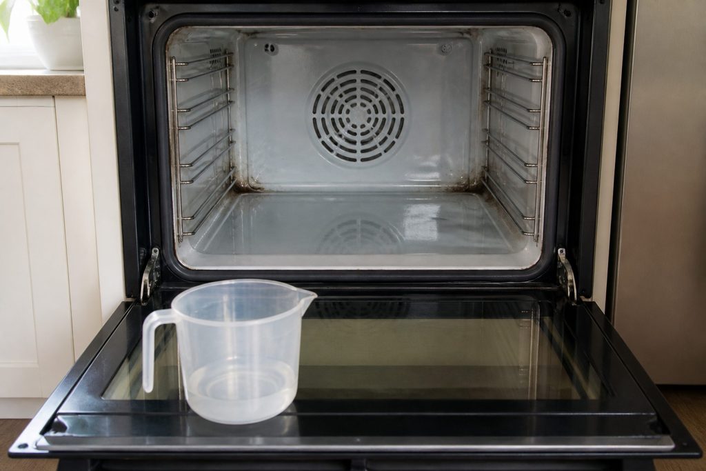 An oven door standing open with a shallow film of water poured across the porcelain cavity floor catching the kitchen light, a plain unmarked measuring jug set down on the open door and softened residue at the back corners.