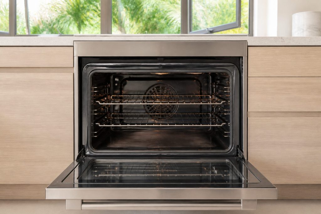 An empty oven cavity with its racks in place and the door standing open, a faint shimmer of heat over the floor of it, a bright South Florida kitchen behind with a window cracked open and greenery outside.
