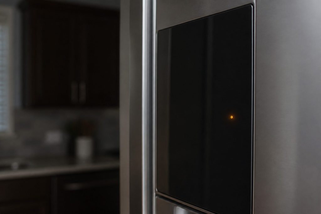 A refrigerator door panel photographed at an angle in a dim kitchen, one small warm indicator lit on dark glass.