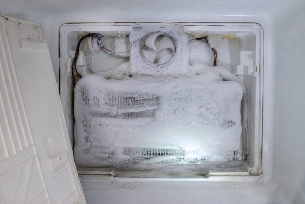 The rear panel of a freezer compartment unscrewed and set aside, the evaporator coil behind it buried in a solid block of white ice so the fins are barely readable, the fan blade above it frozen in place.