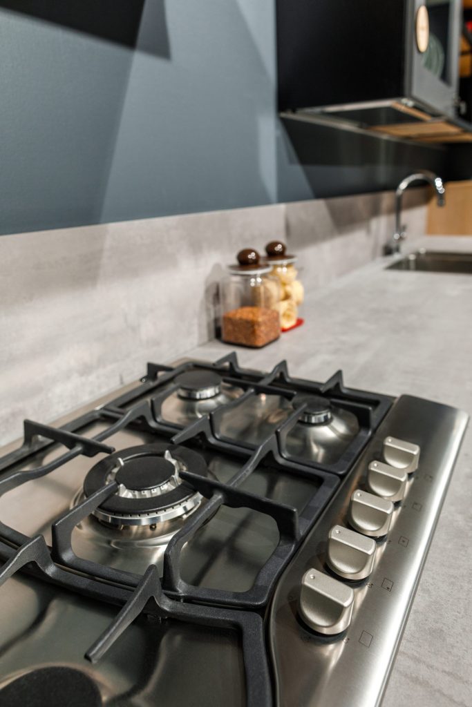 A stainless gas cooktop seen close from one side, four sealed burners with cast grates and a row of knobs along the front edge