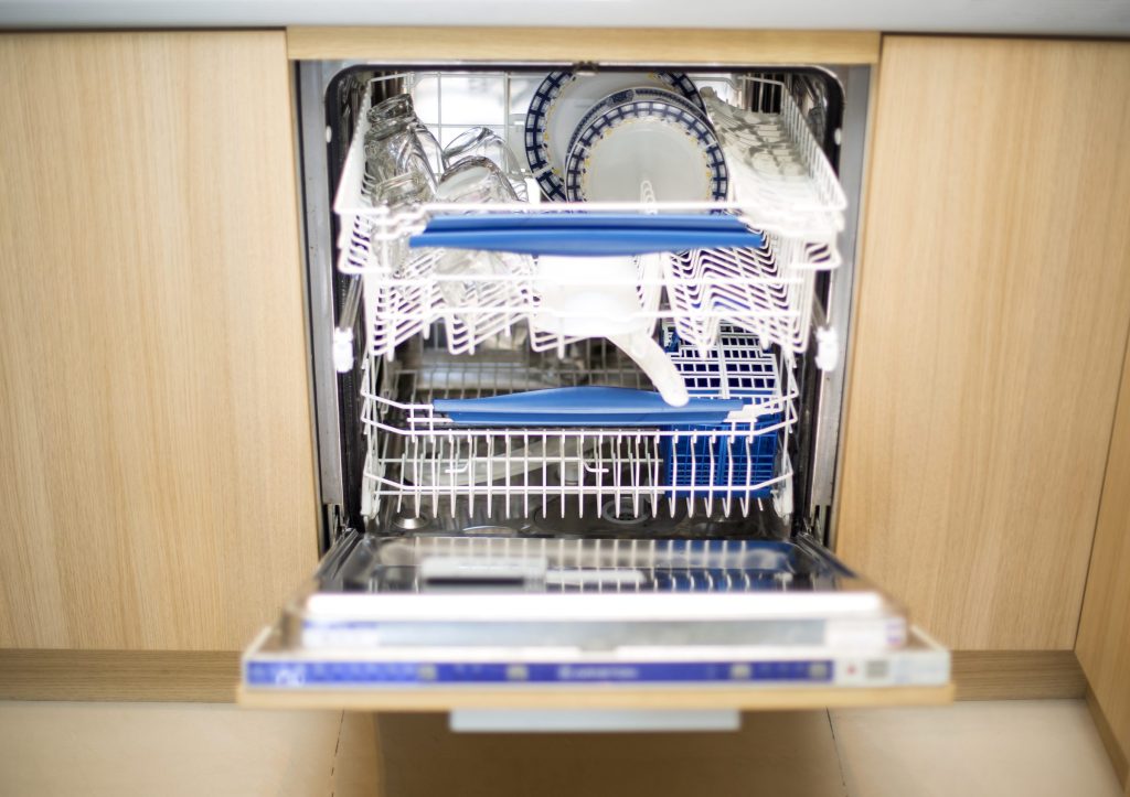 An integrated dishwasher standing open behind a light wood panel, both racks out, a cutlery basket in the lower one, the control strip along the top edge of the door