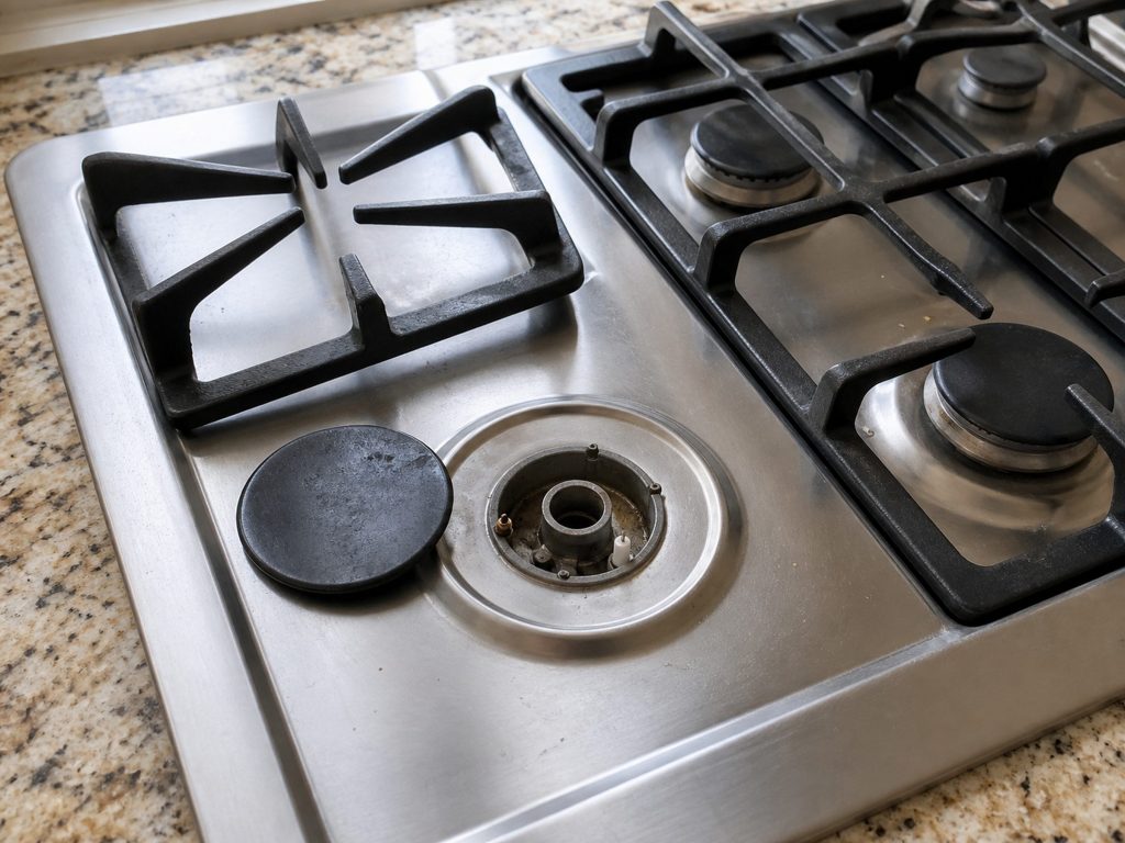 One grate lifted off a gas cooktop and the burner cap tipped beside it, the burner base and the igniter electrode exposed.