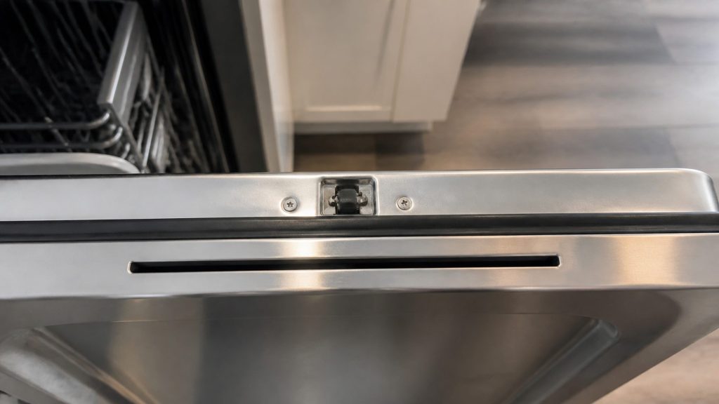 The top edge of an open dishwasher door — the latch, the door seal and the drying vent slot in the inner panel — shot close and slightly from above.