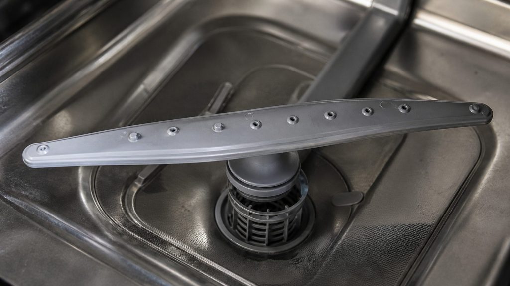 The lower spray arm of a dishwasher photographed close from above, jets set along its length, the sump and filter behind it in shallow focus.