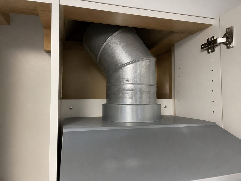 The top of an under-cabinet hood seen from inside the wall cabinet above it — the round duct collar and the first elbow of ducting rising into the void.