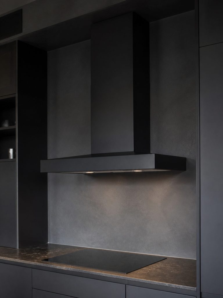 A dark-finish wall-mounted canopy hood in a near-black kitchen, its chimney and canopy reading as one shape against the wall.