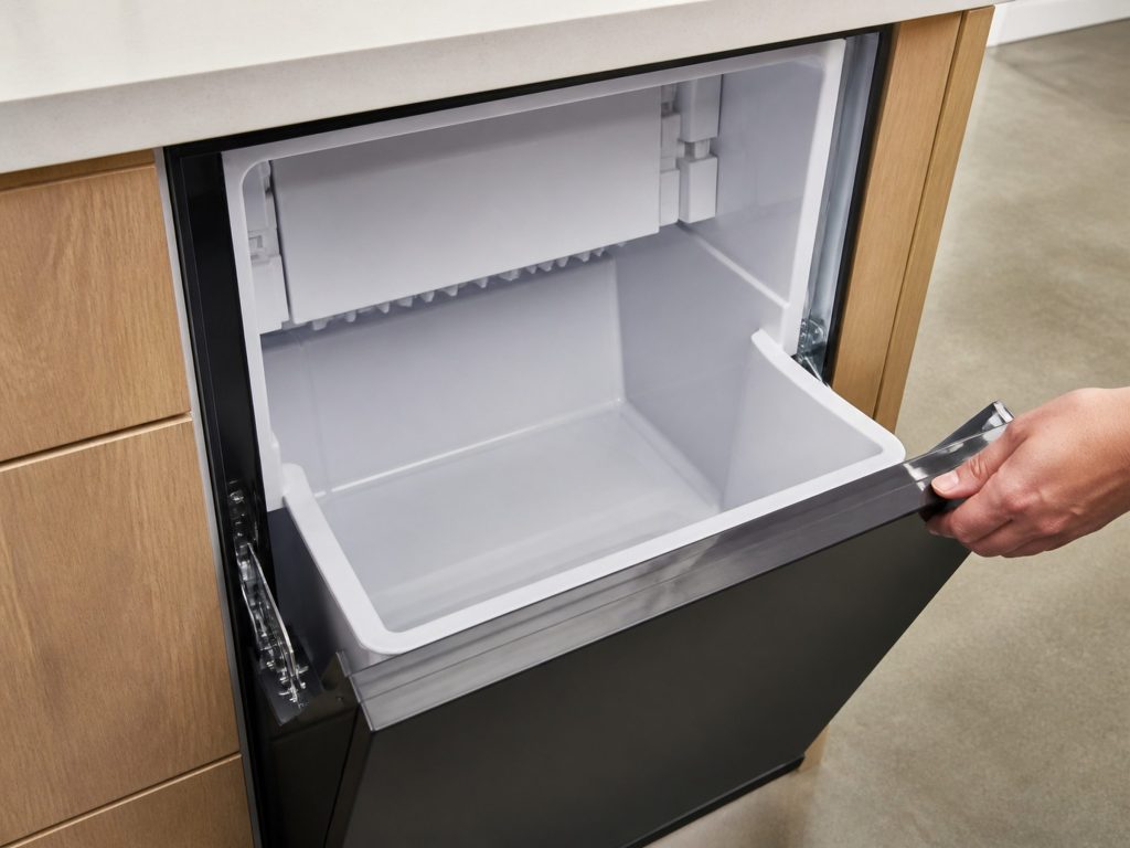 The empty plastic storage bin of an undercounter ice maker after a clean, wiped dry, the door held open by a hand at the edge of the frame.