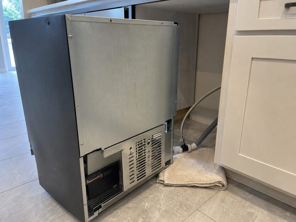 The back of an undercounter ice maker pulled forward on the floor, the water line and the drain pump hose visible behind it, a towel under the joint.