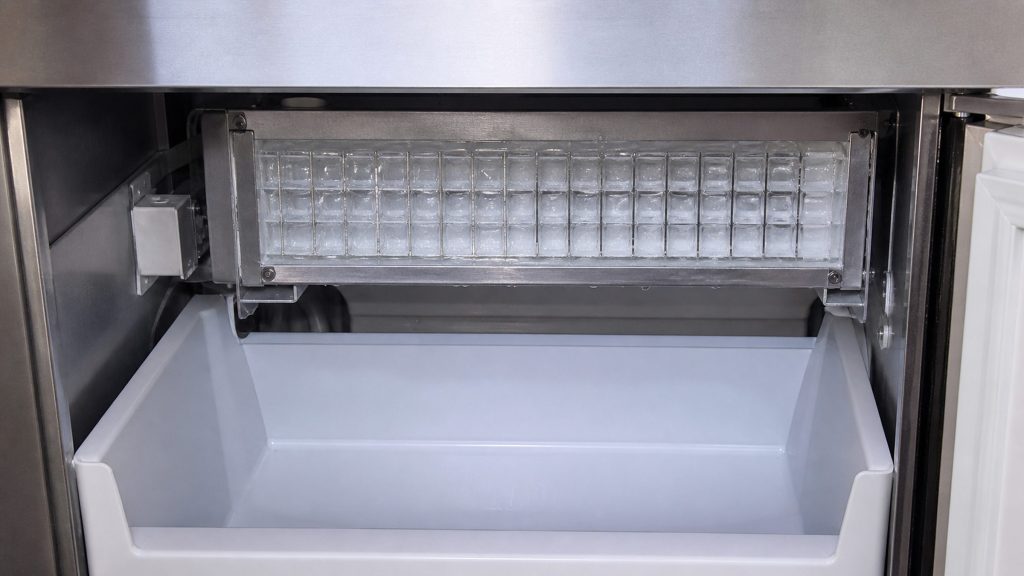 The evaporator plate at the top of an undercounter ice maker's cabinet, a fresh slab of ice formed against its grid, the bin empty below.