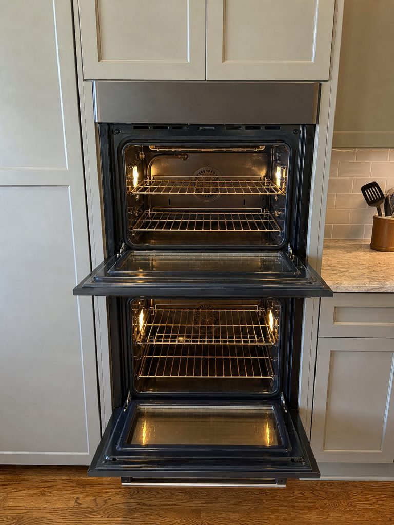 A narrow 24-inch double wall oven built into a tall cabinet, both doors dropped open and both cavities lit, wire racks empty.