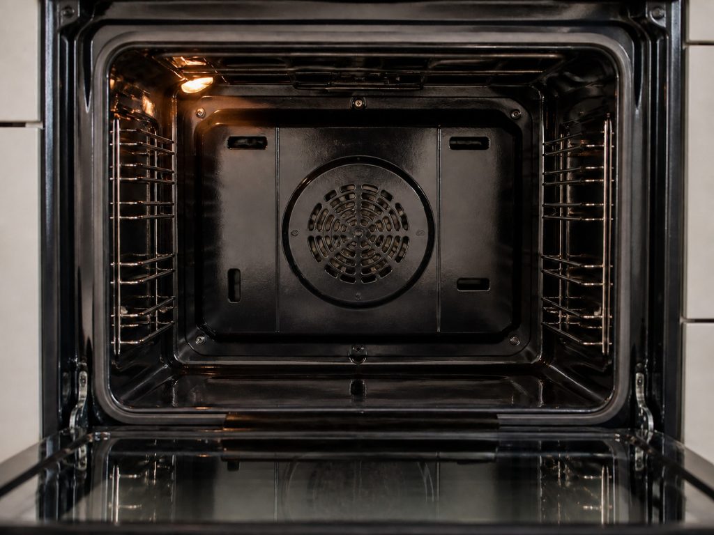 The back wall of an oven cavity with the convection fan cover and its circular grille in the center, the door open and the cavity light on.
