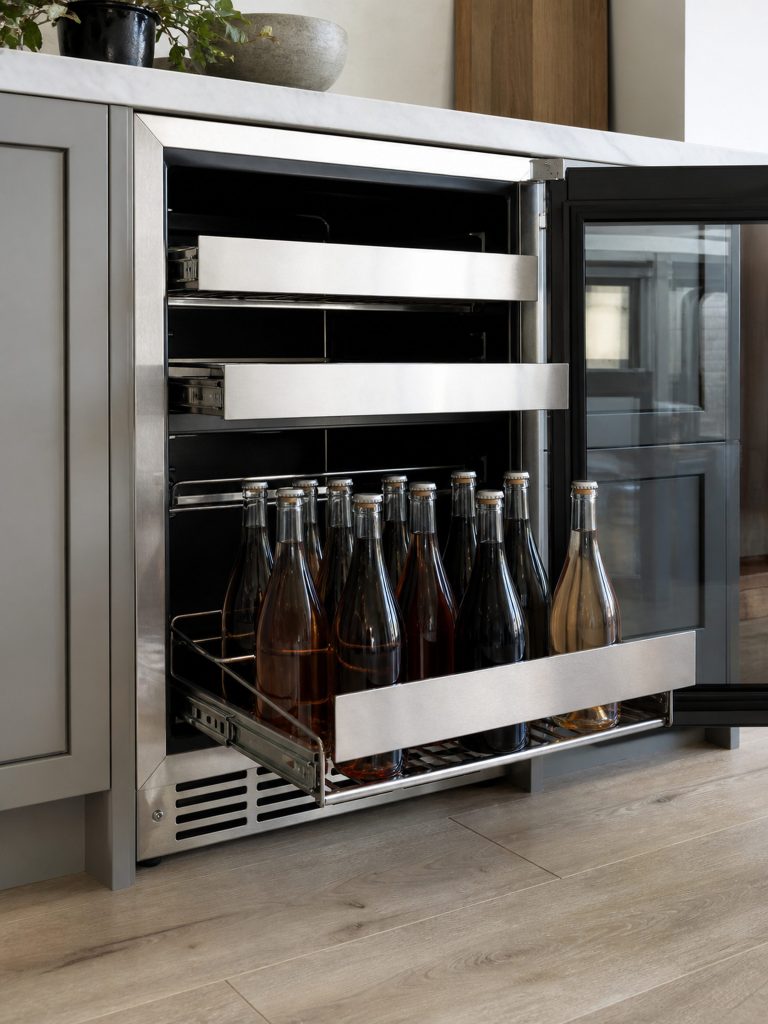 A stainless undercounter beverage center with metal-front racks pulled part way out, bottles standing upright on the lowest shelf.