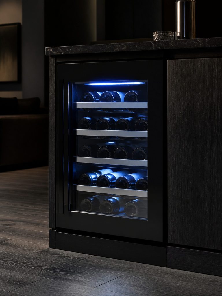 A 24-inch undercounter wine cellar with a glass door, the interior lit blue-white, bottles lying on metal-front racks in a dark room.