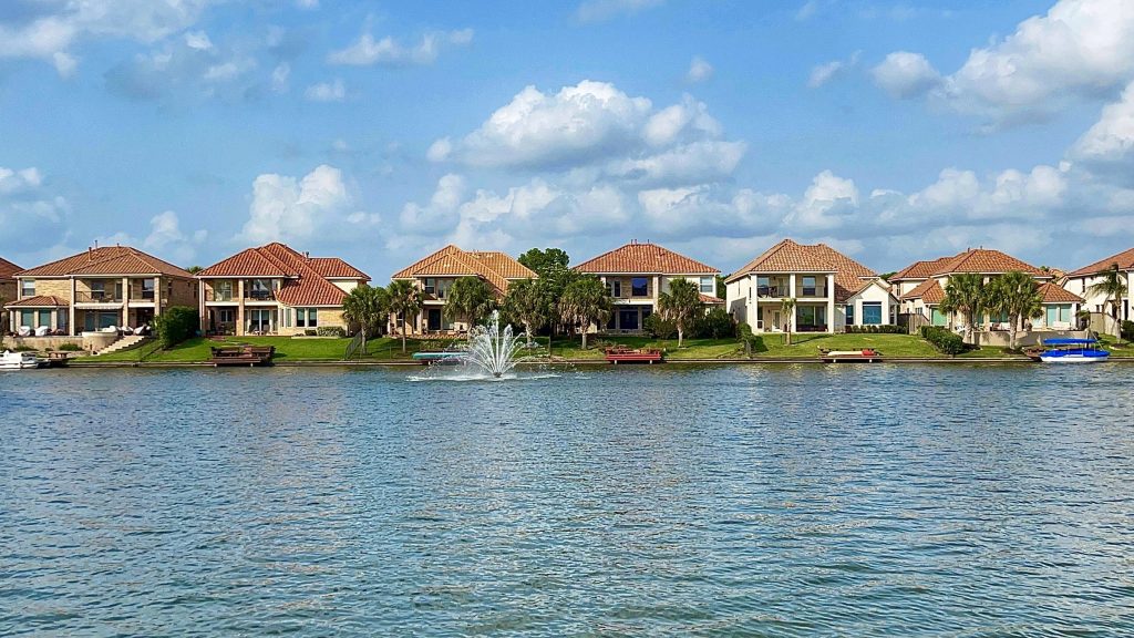 Two-story waterfront houses with terracotta roofs along a lagoon, a fountain playing in the middle