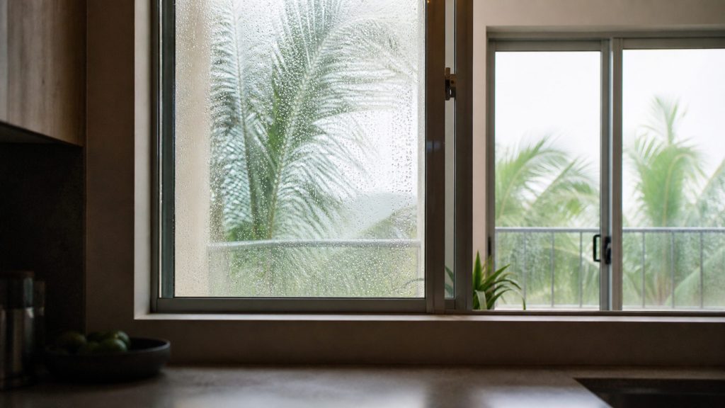 A kitchen window with sliding glass beyond it, condensation beading on the outside of the glass, palm fronds and bright hazy sky past the terrace.