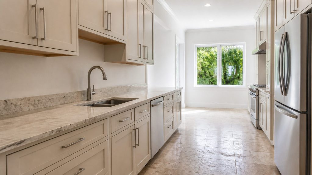 A wide South Florida kitchen photographed along the counter run, pale cabinetry and a light stone worktop, stainless appliance fronts set back and partly cropped at the right edge, bright even daylight from a window at the far end with green foliage outside.