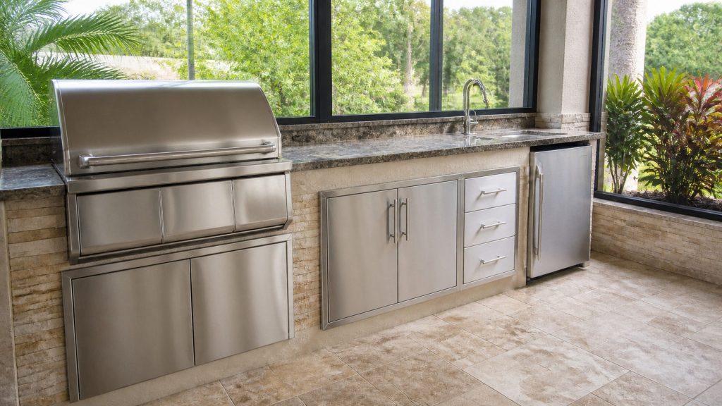 A covered outdoor kitchen counter on a terrace, stainless doors under a stone top, screened enclosure and planting beyond, bright shade.