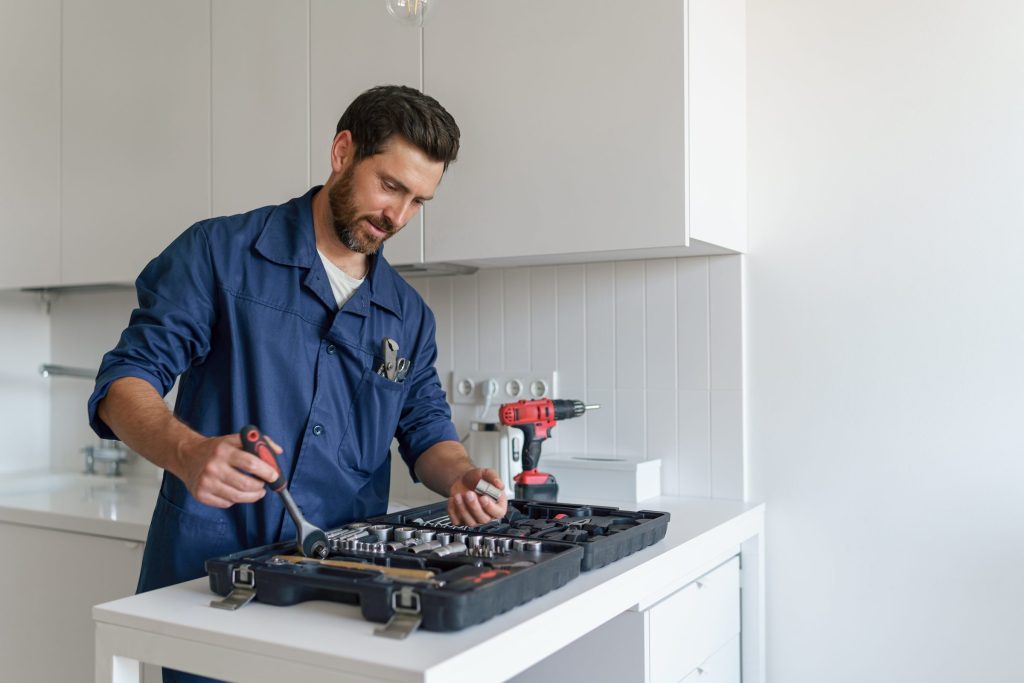 A repair technician in workwear laying out a socket set on a kitchen counter beside a cordless driver