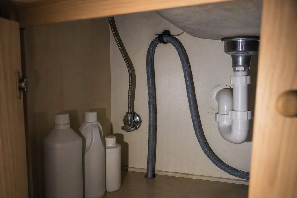 The inside of a base cabinet under a kitchen sink: a shut-off valve, a braided supply line and a corrugated drain hose looping up, cleaning bottles pushed to one side.