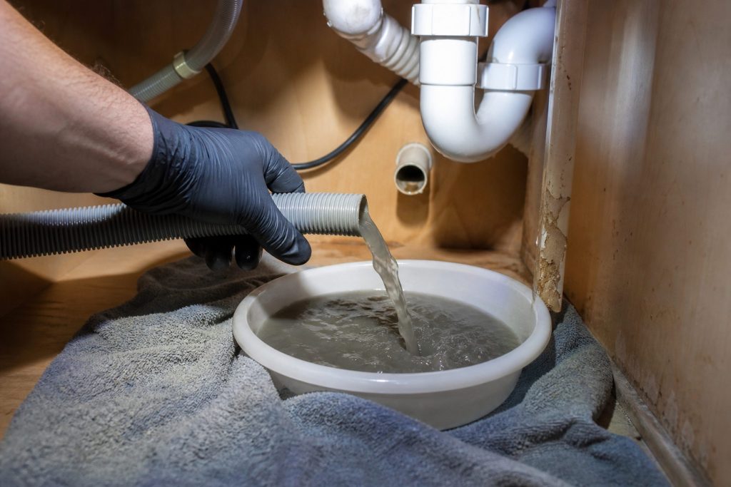 A corrugated dishwasher drain hose disconnected from its spigot inside a base cabinet under a kitchen sink and held down over a shallow plastic bowl, grey water running out of the end of it, one gloved hand steadying the hose and a folded towel spread under the bowl.