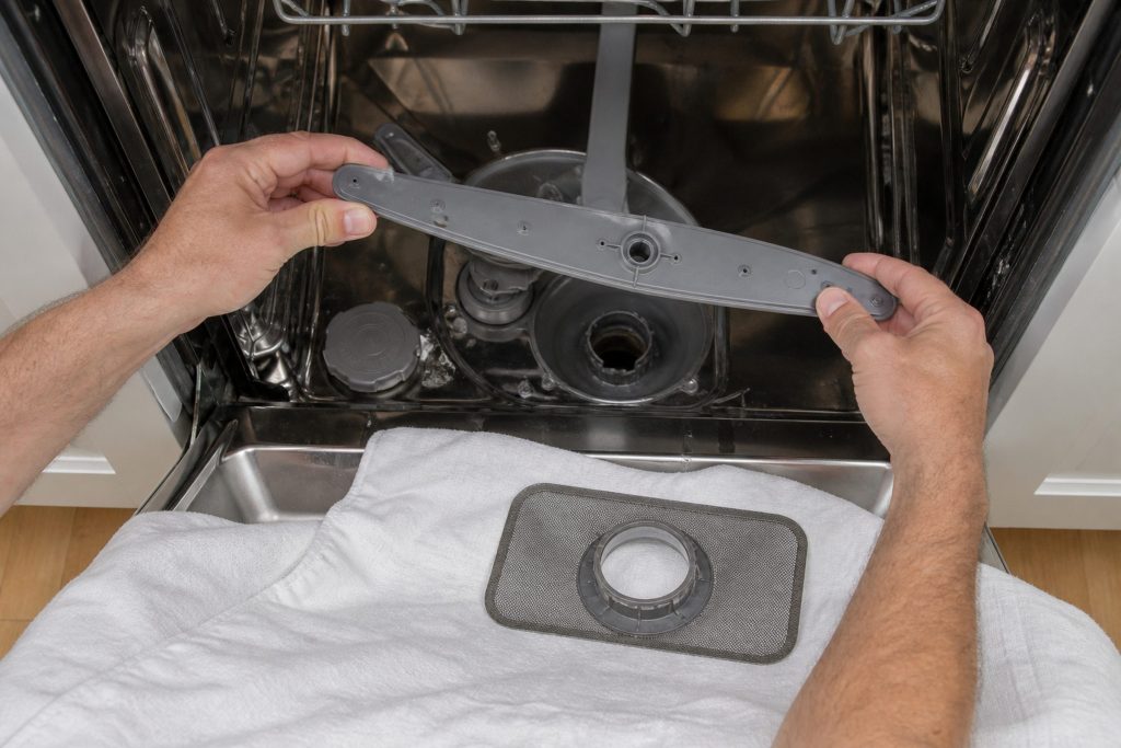 The lower spray arm of a dishwasher unclipped and lifted a few inches clear of its hub by two hands, the fine mesh screen already lying on a cloth on the open door beside it, the empty sump visible below.