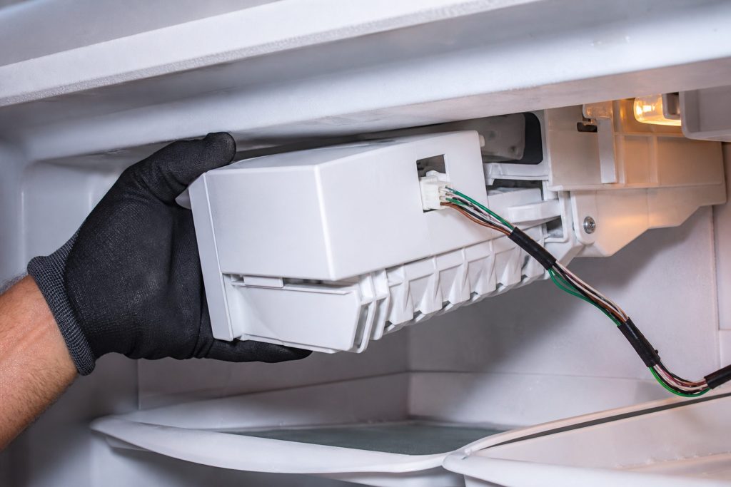 A gloved hand easing an ice maker module out of the top of a freezer compartment, its wiring harness still attached.