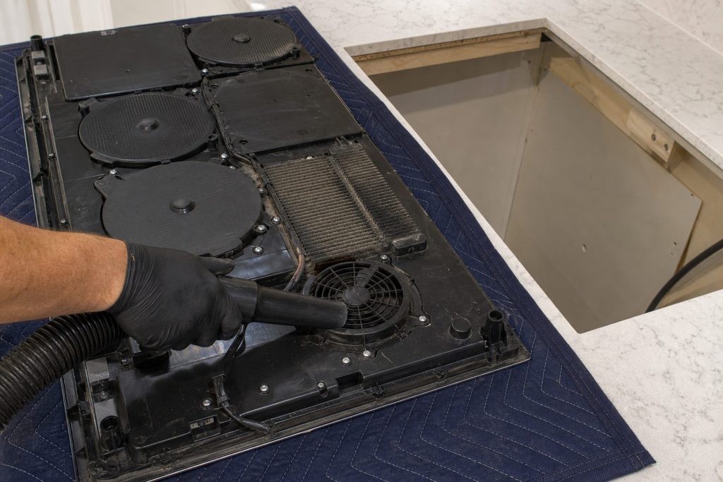 An induction cooktop lifted clear of the counter and laid on a blanket beside its opening, the underside toward the camera showing the finned electronics module, the cooling fan and the coil housings, a gloved hand holding a vacuum nozzle to the fan intake and dust visible on the fins.