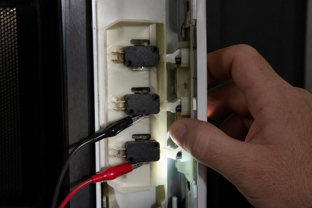 The latch board of an opened microwave door frame with its three small interlock switches in a row, meter clip leads hooked onto two of them and a hand working the latch hooks in and out.
