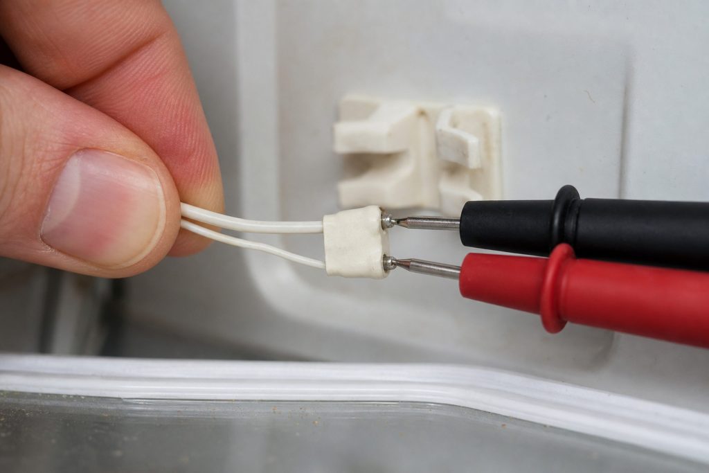 A small thermistor unclipped from its bracket on an interior refrigerator wall and drawn out on its two-wire lead into the cabinet, held between finger and thumb, with a pair of meter probes touching its terminals from the other side of the frame.