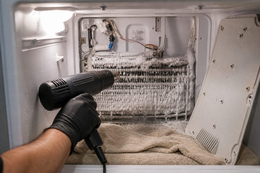 The rear cover of a freezer compartment unscrewed and set aside, the evaporator coil behind it thick with white ice, a gloved hand playing a heat gun across the fins at an angle with a towel packed along the bottom of the compartment to catch the melt.