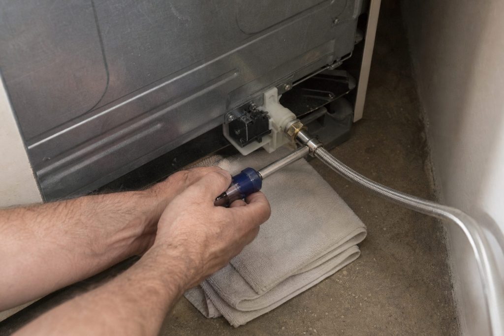 Hands with a nut driver working on the water inlet valve on the lower back of a refrigerator that has been pulled out from the wall, the braided supply line arriving from the right and a folded towel under the fitting.