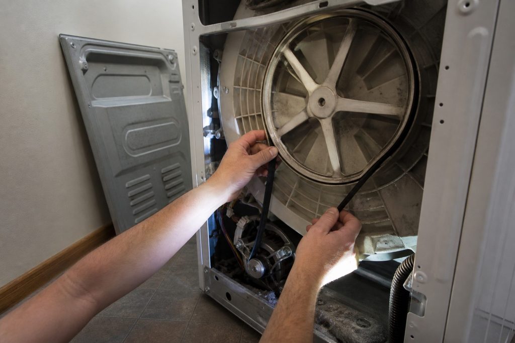 The rear panel of a washing machine removed and leaning against the wall, the drive belt looped around the motor pulley and the large drum pulley behind it, two hands walking the belt onto the pulley.