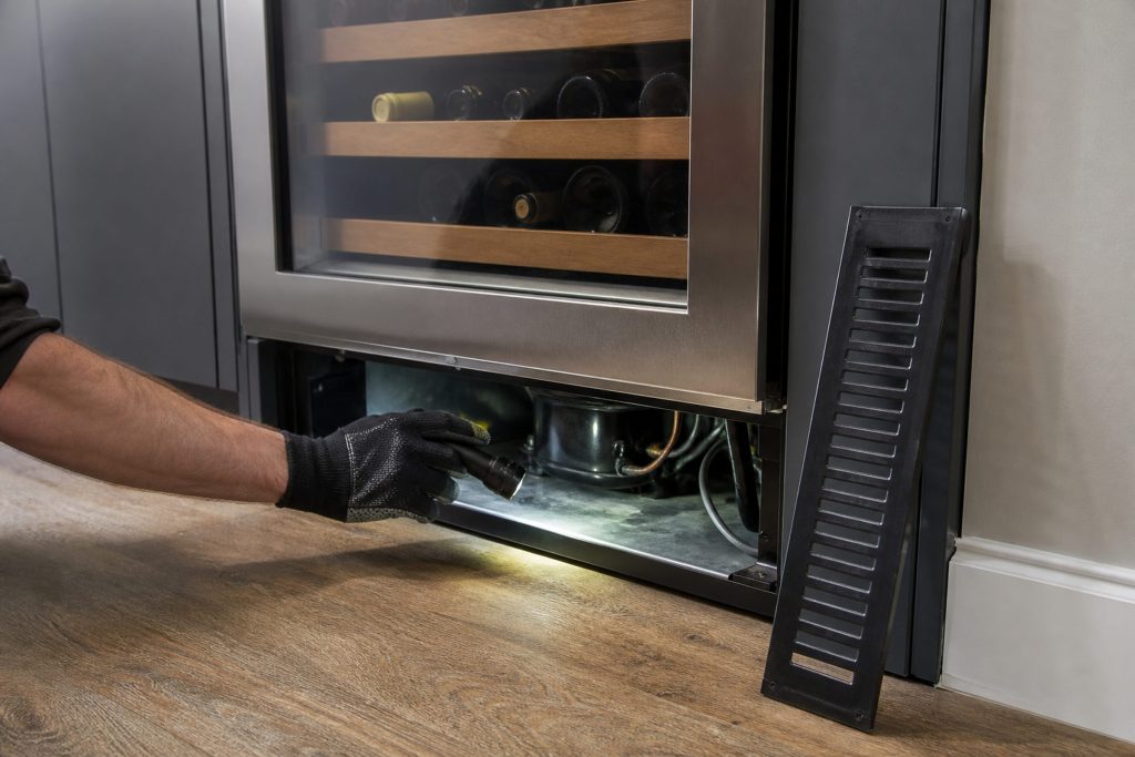 A gloved hand reaching behind the toe-kick grille of an undercounter wine cabinet with a torch, the grille propped against the cabinet beside it.