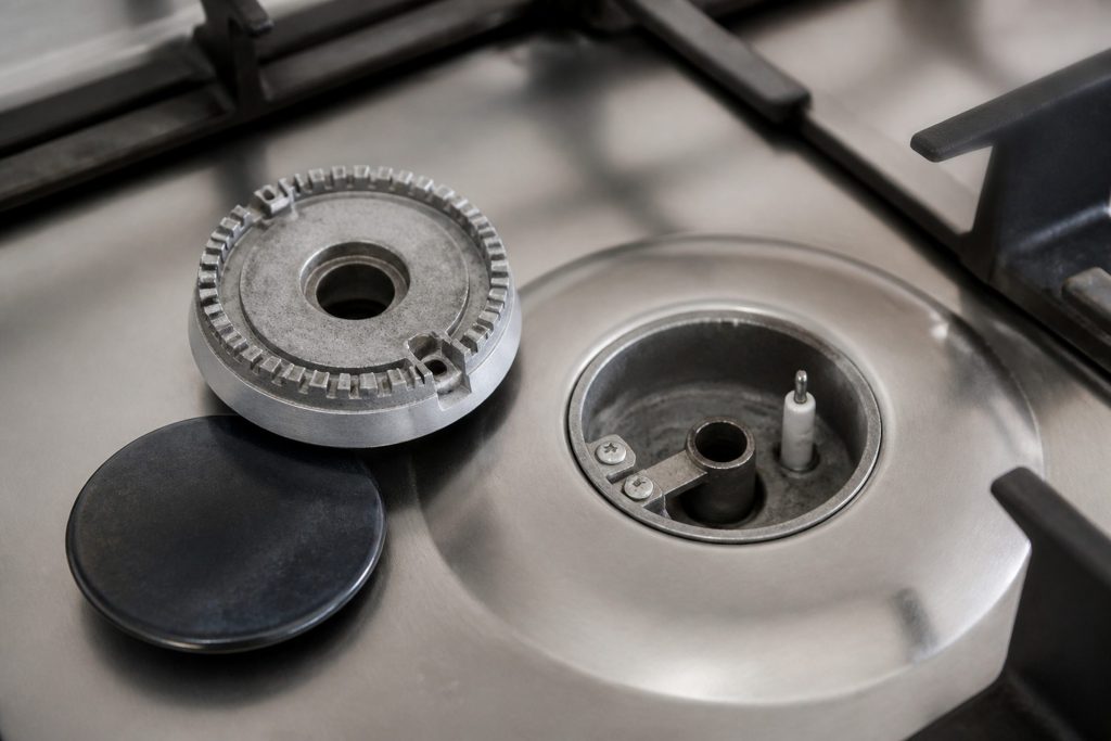 A gas burner head and cap lifted off and set on the cooktop beside its empty well, the igniter electrode visible in the well.