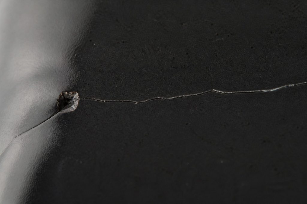 A crack running across black ceramic from a small chipped point, lit from the side so the line catches the light.