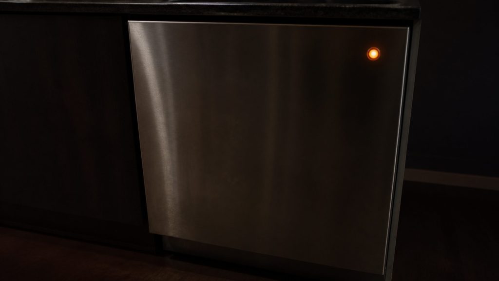 A stainless dishwasher door at counter height in a dim kitchen, one small round indicator glowing warm orange on the otherwise blank steel.