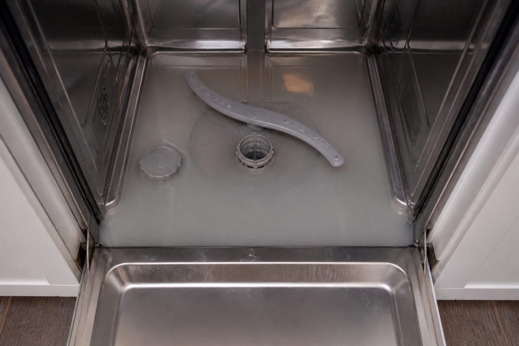 Looking down into the bottom of an open dishwasher tub with two inches of grey standing water covering the floor of it, the lower rack lifted out and set aside.
