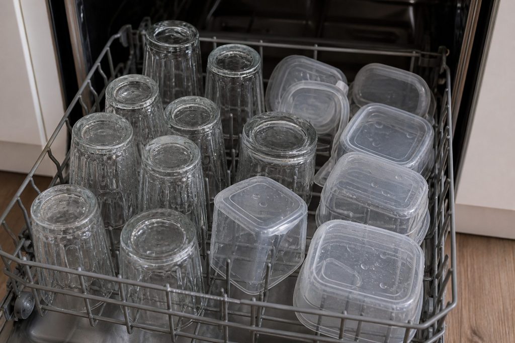 An open dishwasher rack of upturned glasses and plastic containers with water still pooled in every hollow, steam gone, close from slightly above.