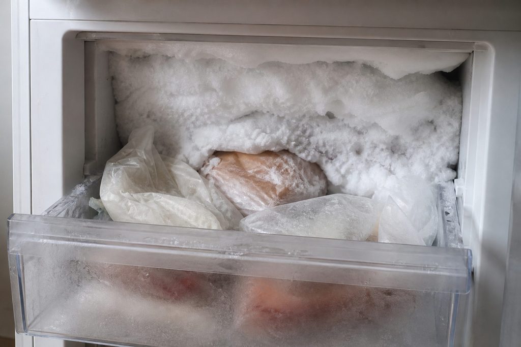 The back wall of a freezer compartment carrying a thick white ridge of frost, packages half buried in it, the drawer pulled forward.