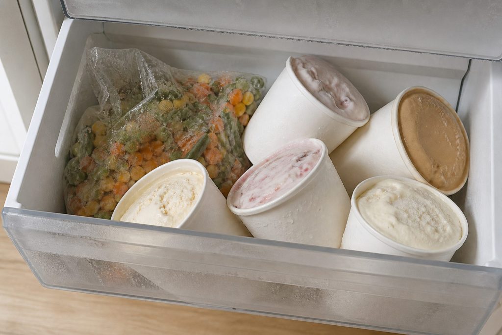 An open freezer drawer with tubs of ice cream slumped soft and a bag of vegetables collapsed, moisture on the packaging.