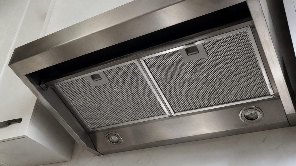 Looking up at the underside of a stainless range hood, metal mesh filters in place, the lights off and the cavity dark.