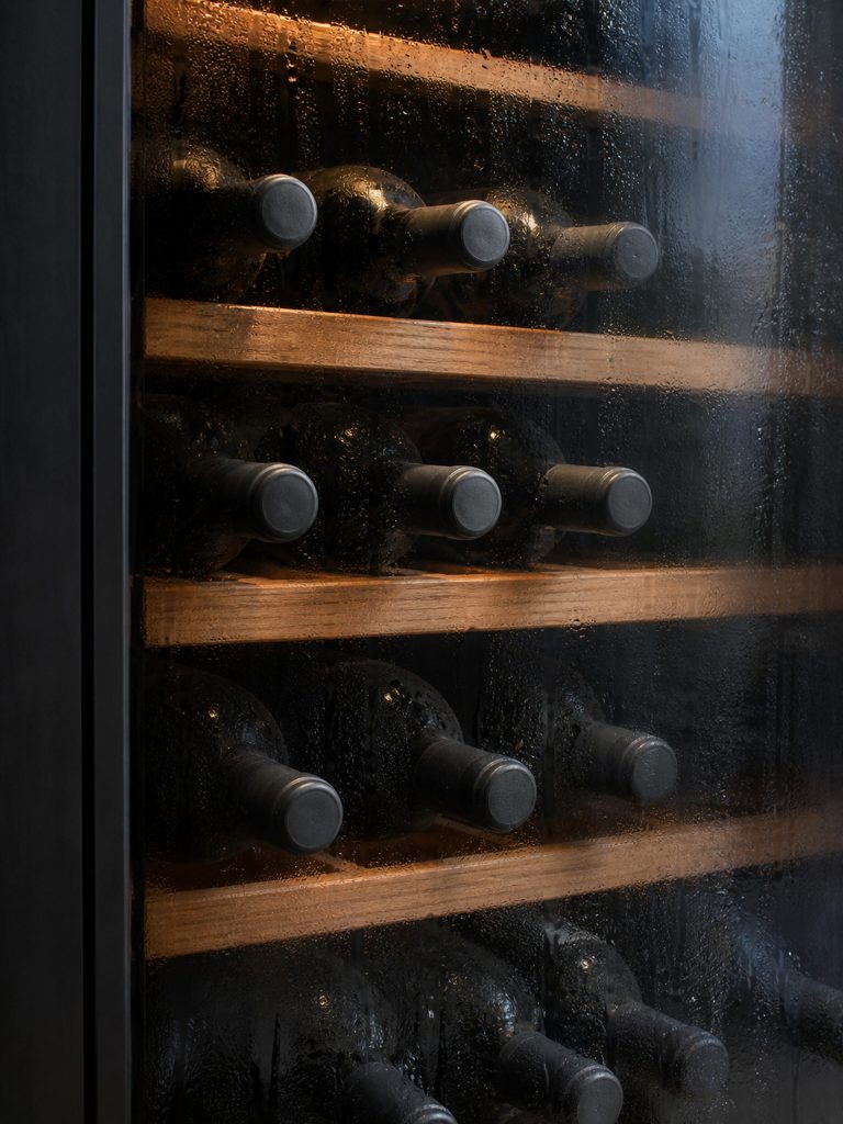 Wine bottles lying on wooden shelves behind smoked glass, the inside of the glass fogged with condensation.